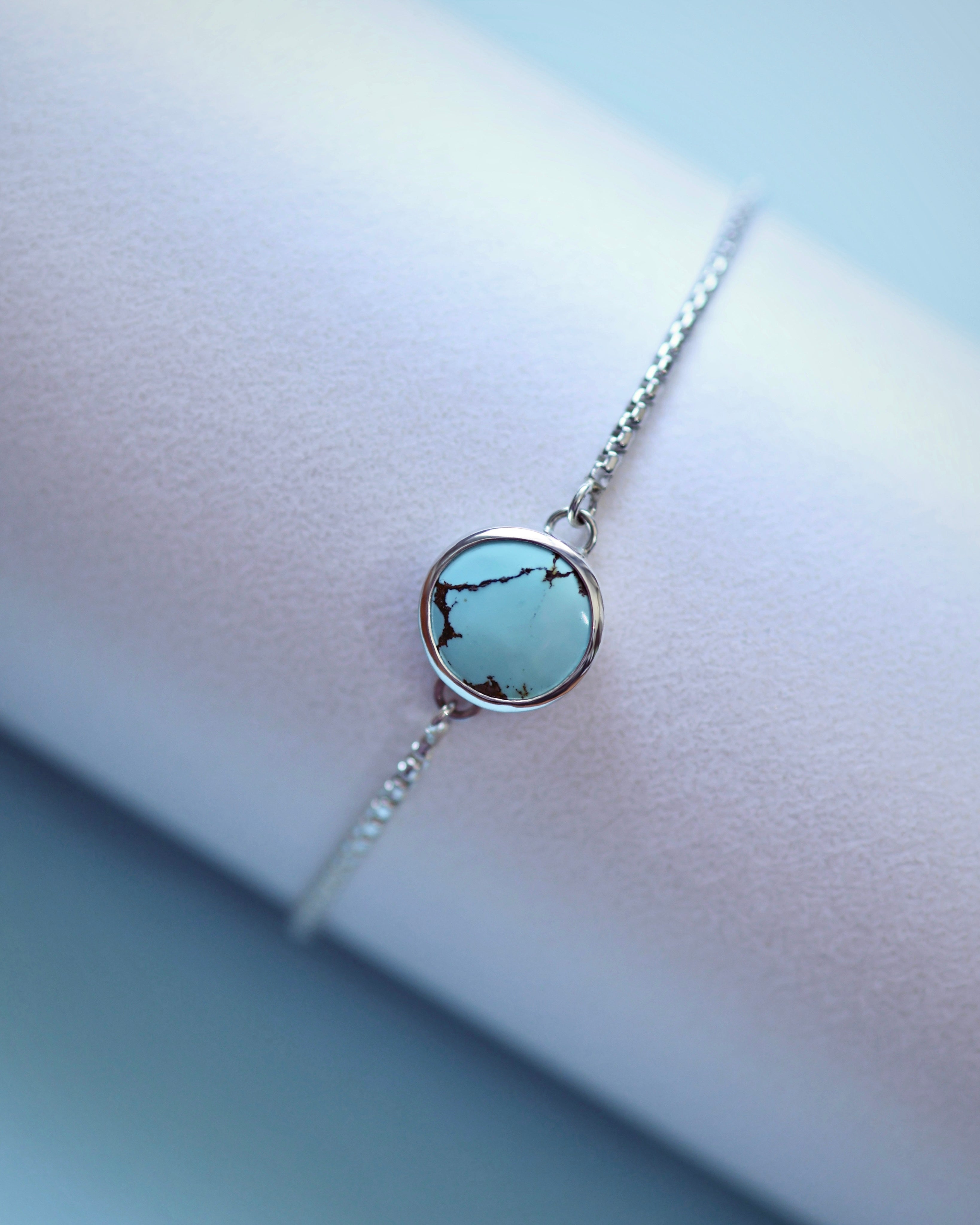 The Cloud Mountain Turquoise Bracelet No. 1 by Lilac Sky Studio features a round turquoise stone with dark veining set in sterling silver and an adjustable chain, elegantly displayed on a pale surface.