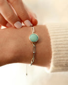 A hand wears Lilac Sky Studio's Treasure Mountain Turquoise Bracelet No. 1, featuring a round turquoise stone and adjustable sterling silver cable chain, shown against a cream-colored sweater.