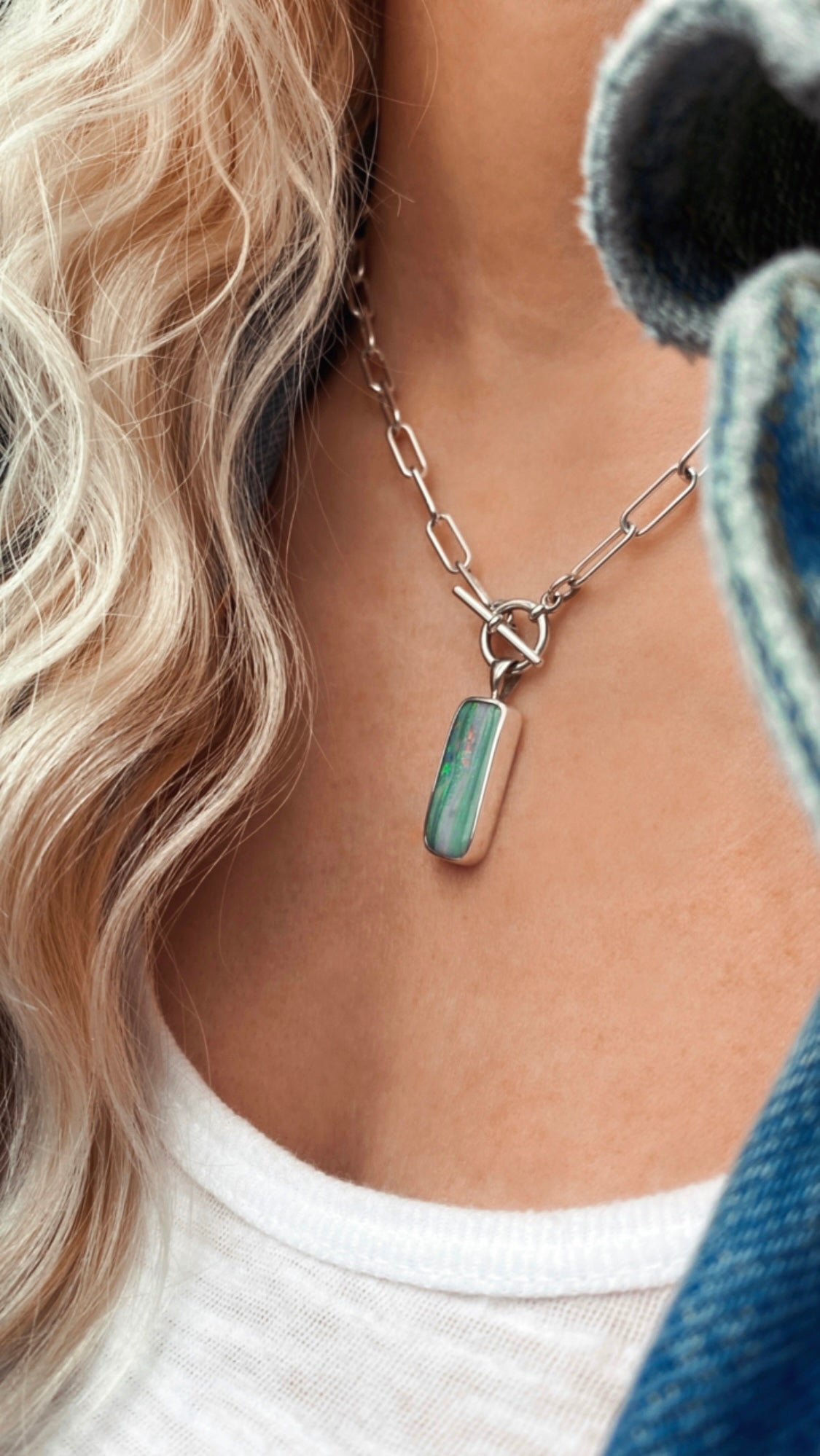 Close-up of a person wearing a handcrafted silver chain necklace with a rectangular green and pink stone pendant, white top, and light curly hair visible.