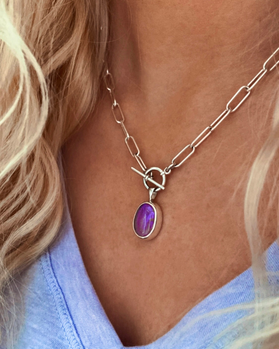 A person wears the Lilac Sky Studio Crystal Opal Necklace, a sterling silver chain with a toggle clasp and purple oval opal pendant, styled with a light lavender V-neck shirt.