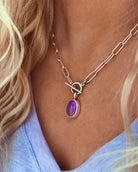 A person wears the Lilac Sky Studio Crystal Opal Necklace, a sterling silver chain with a toggle clasp and purple oval opal pendant, styled with a light lavender V-neck shirt.