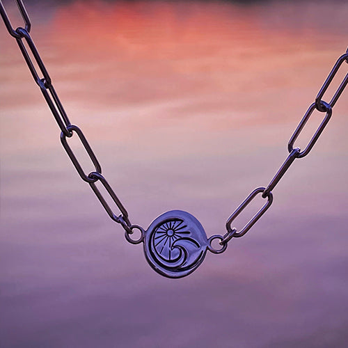 A chain necklace with a circular pendant featuring an engraved abstract design, handcrafted and displayed against a blurred sunset sky.