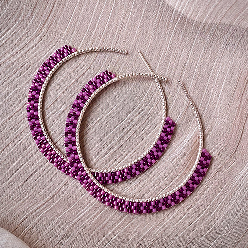 A pair of large hoop earrings with alternating purple and silver beads, this elegant jewelry piece is displayed on a textured light pink fabric background.