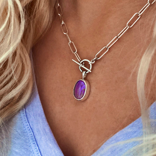 A close-up of a person wearing gemstone jewelry: a silver chain necklace with a purple oval gemstone pendant, paired with a light blue V-neck top.