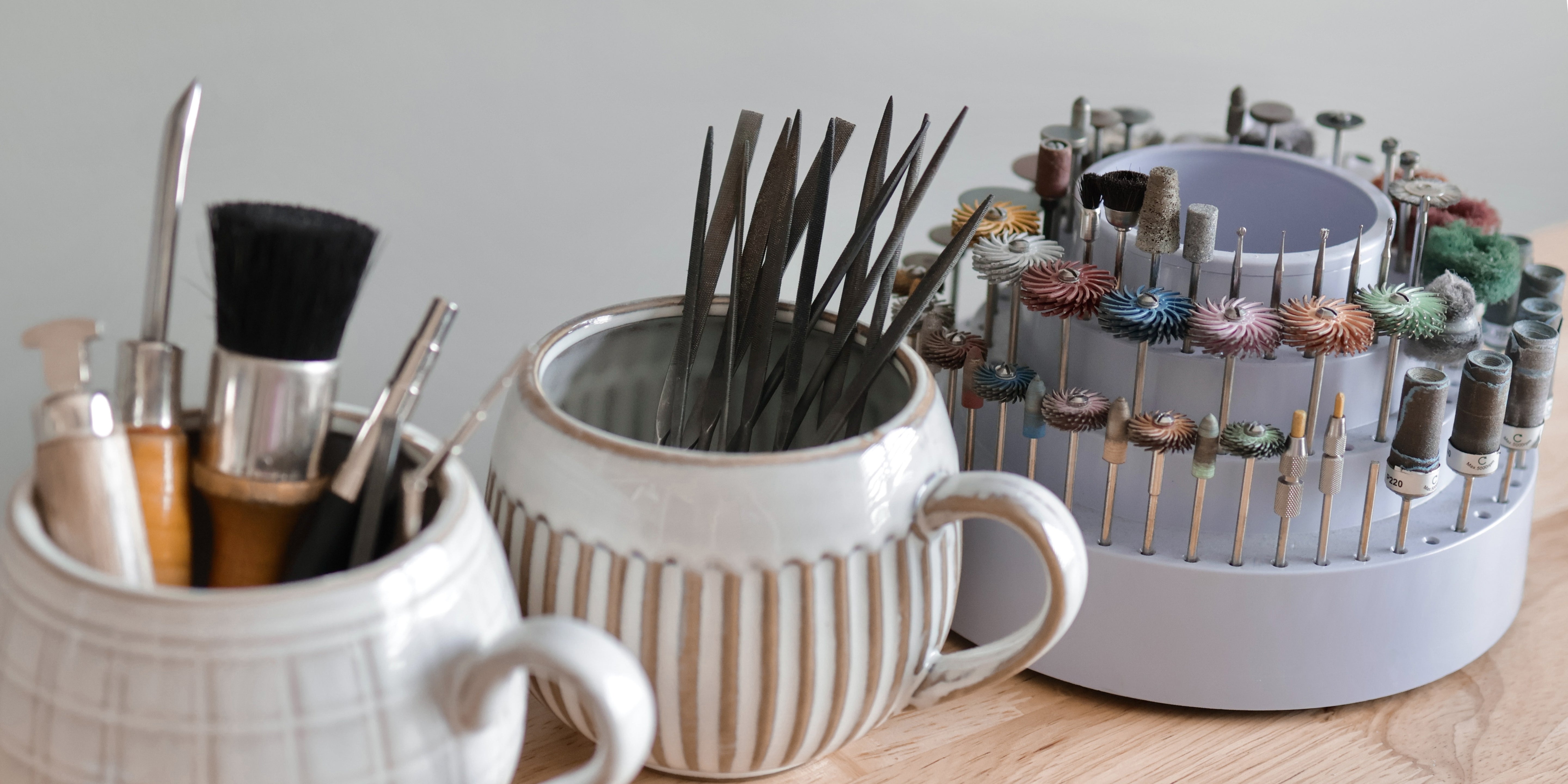 Various handcrafted jewelry-making tools, files, brushes, and rotary tool attachments are organized in ceramic mugs and a round holder on a wooden surface, ready for creating unique gold pieces.
