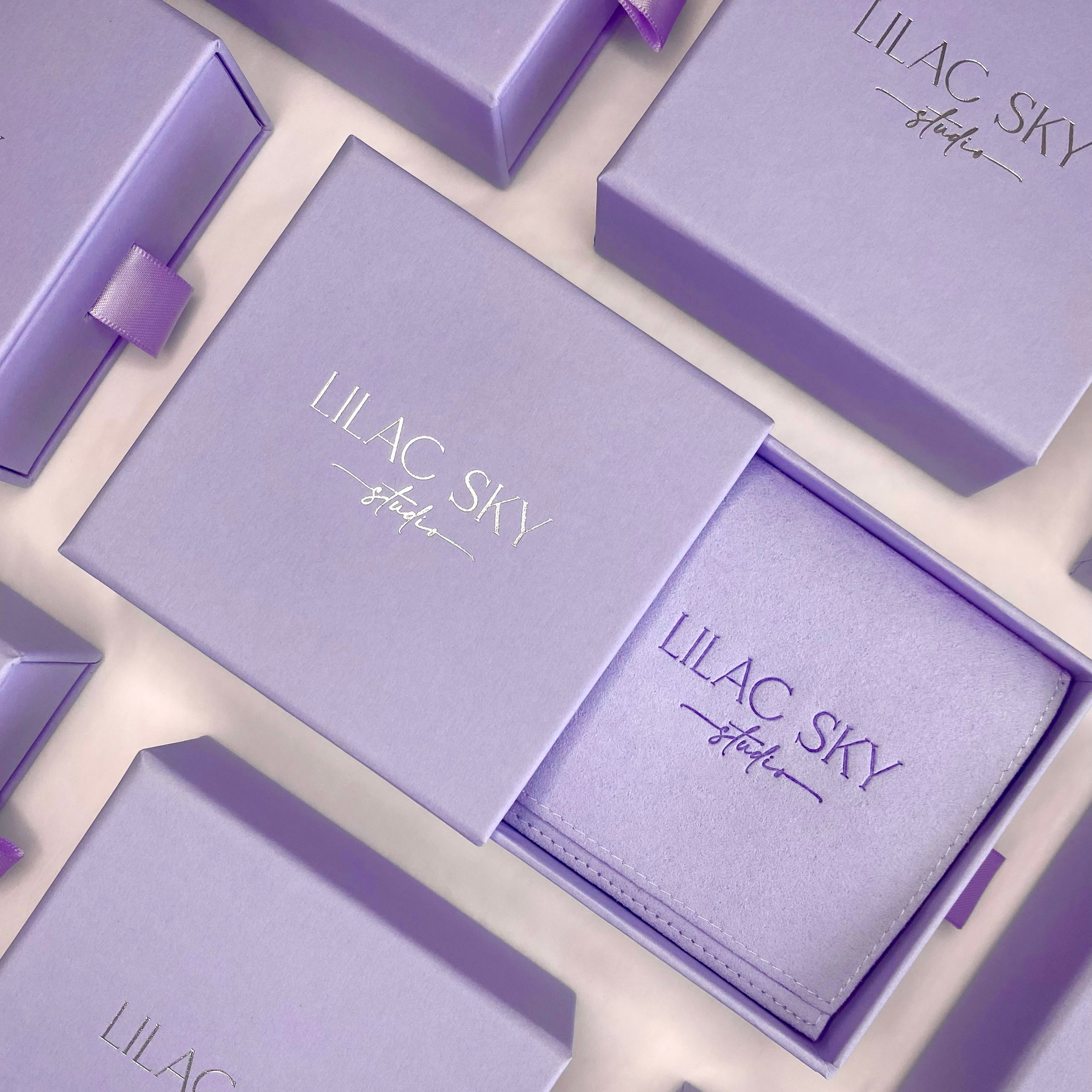 A lilac-colored wallet in a matching gift box, both labeled "LILAC SKY store," is surrounded by similar boxes with ribbon pulls, perfect for pairing with handcrafted jewelry or gold accessories.