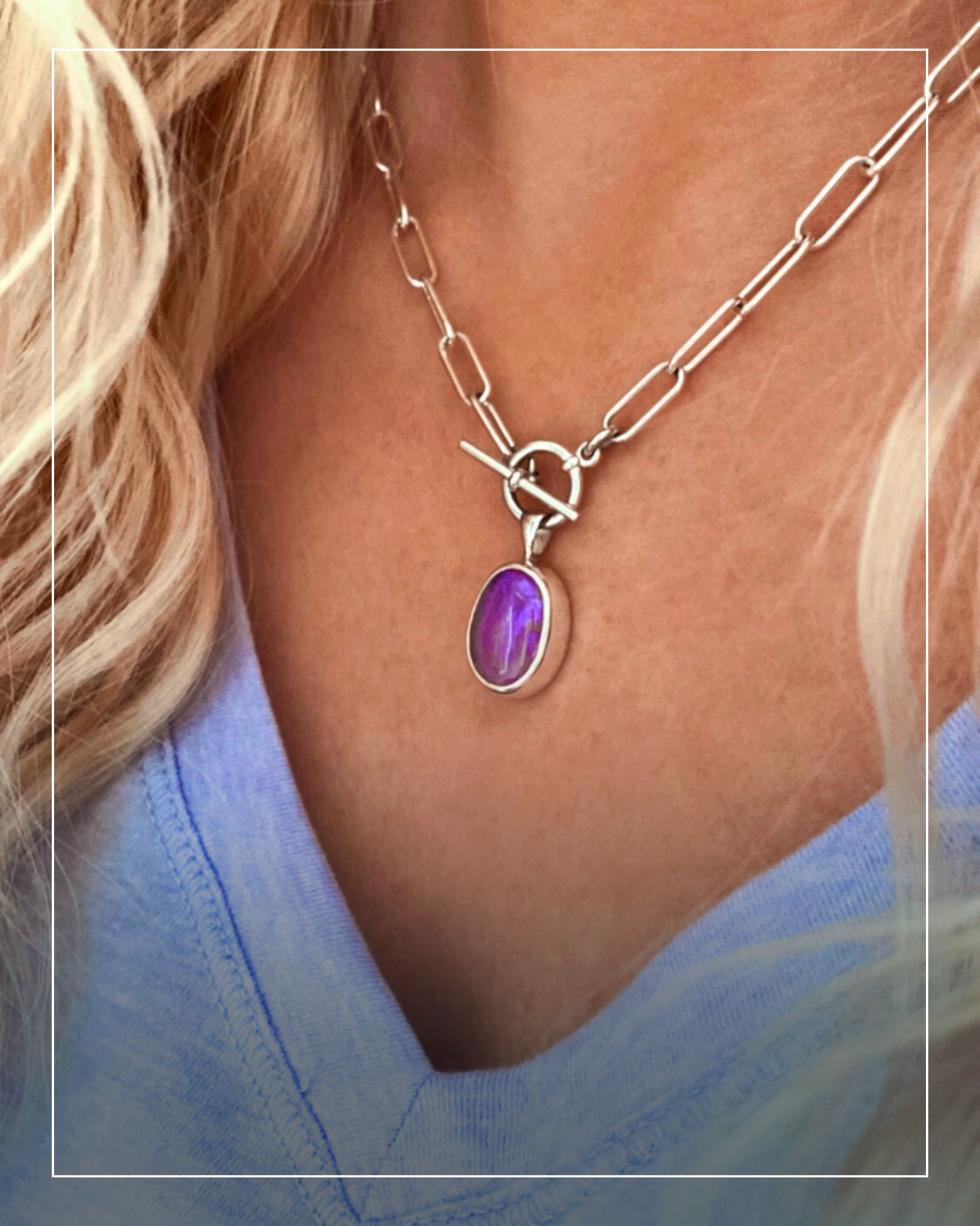 A person wearing a handcrafted silver chain necklace with an oval purple gemstone pendant, shown against a light blue v-neck shirt.