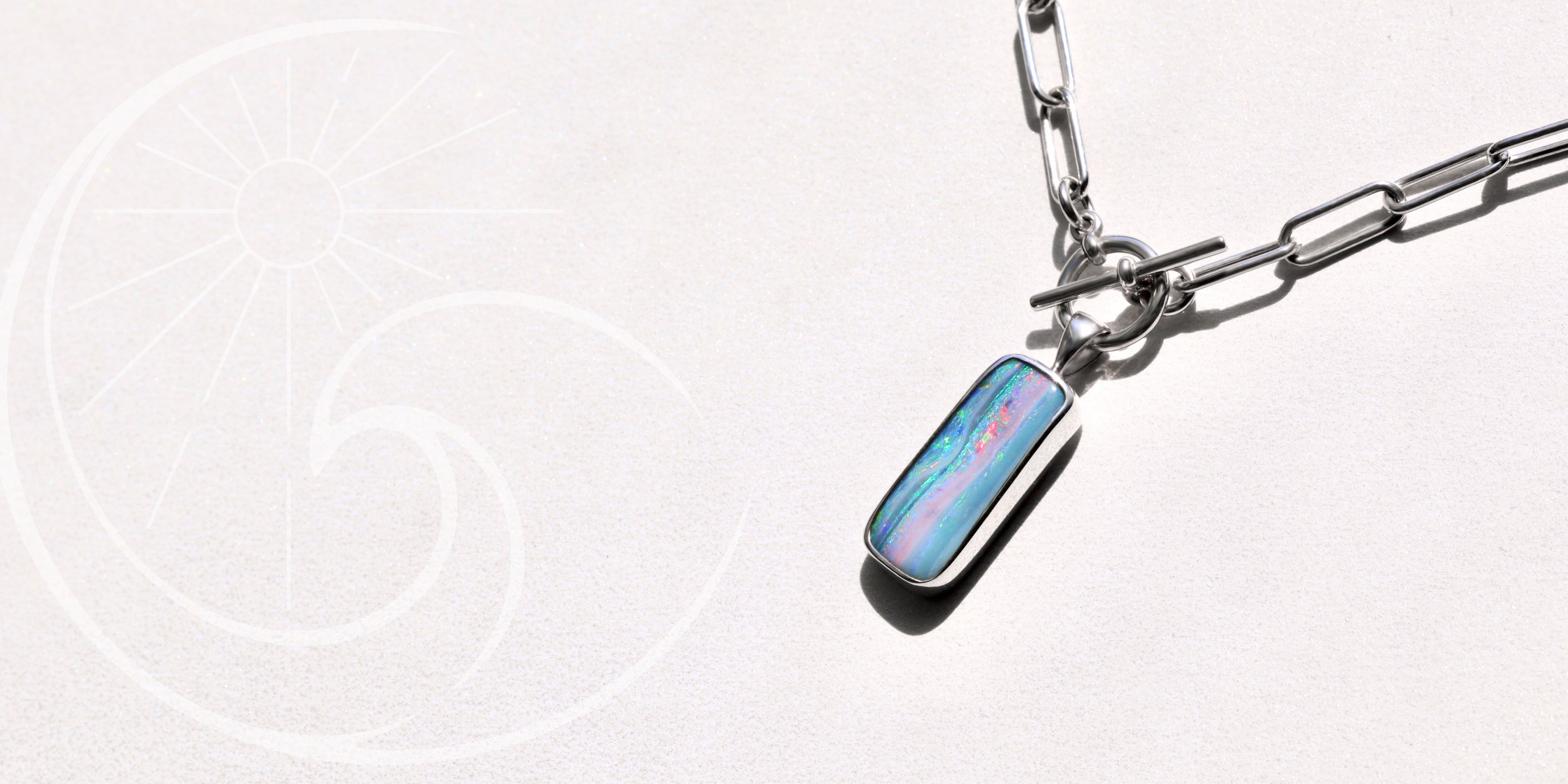 A silver chain necklace with a handcrafted rectangular opal gemstone pendant is displayed on a light background with a subtle sun and wave design.