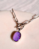 The Lilac Sky Studio Crystal Opal Necklace, featuring a sterling silver chain and an oval purple crystal opal pendant, rests on a light textured surface.