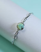 The Lilac Sky Studio Treasure Mountain Turquoise Bracelet No.3 features a round turquoise stone with dark veins on a sterling silver cable chain, displayed on a white cylinder against a light blue background.