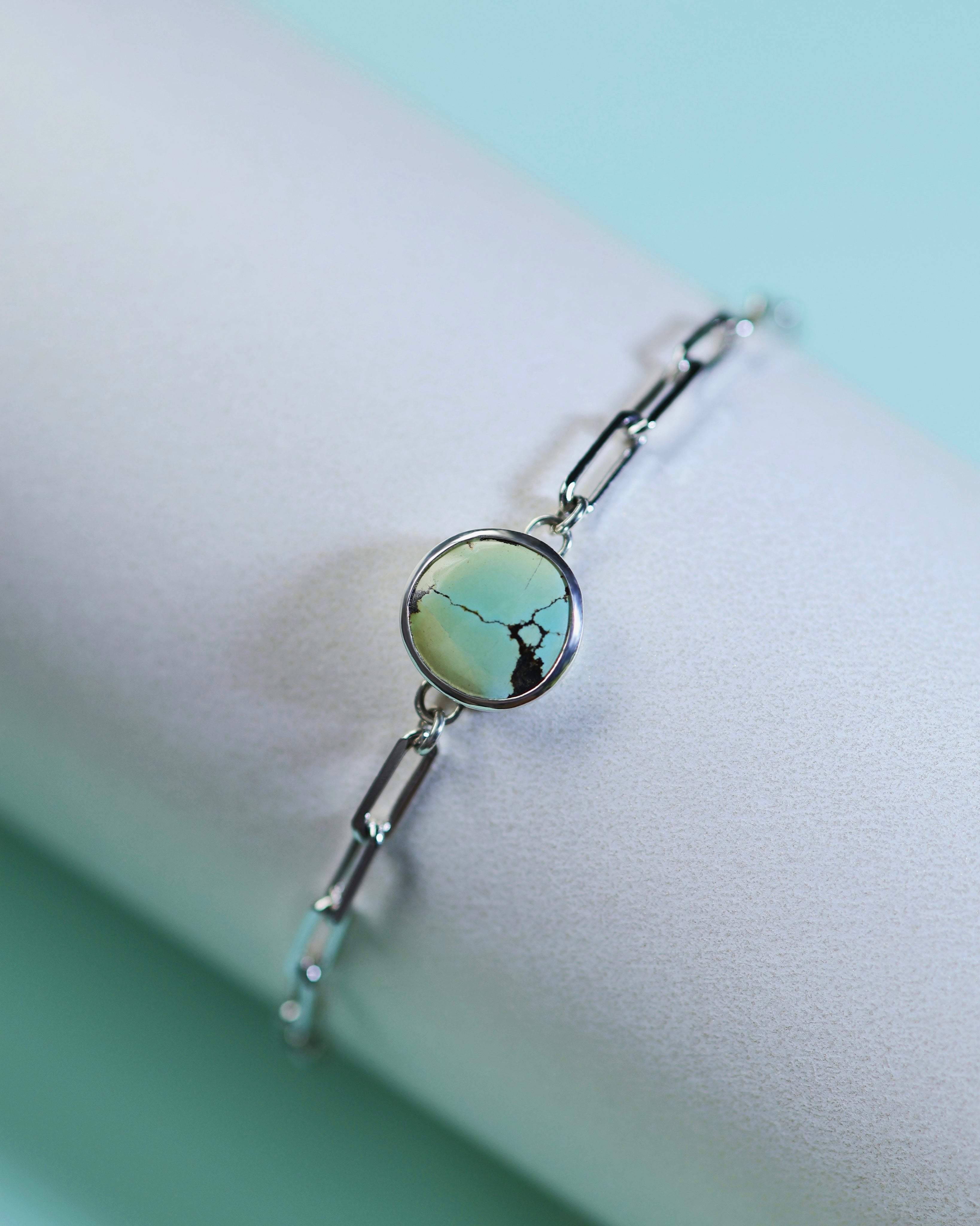 The Lilac Sky Studio Treasure Mountain Turquoise Bracelet No.3 features a round turquoise stone with dark veins on a sterling silver cable chain, displayed on a white cylinder against a light blue background.