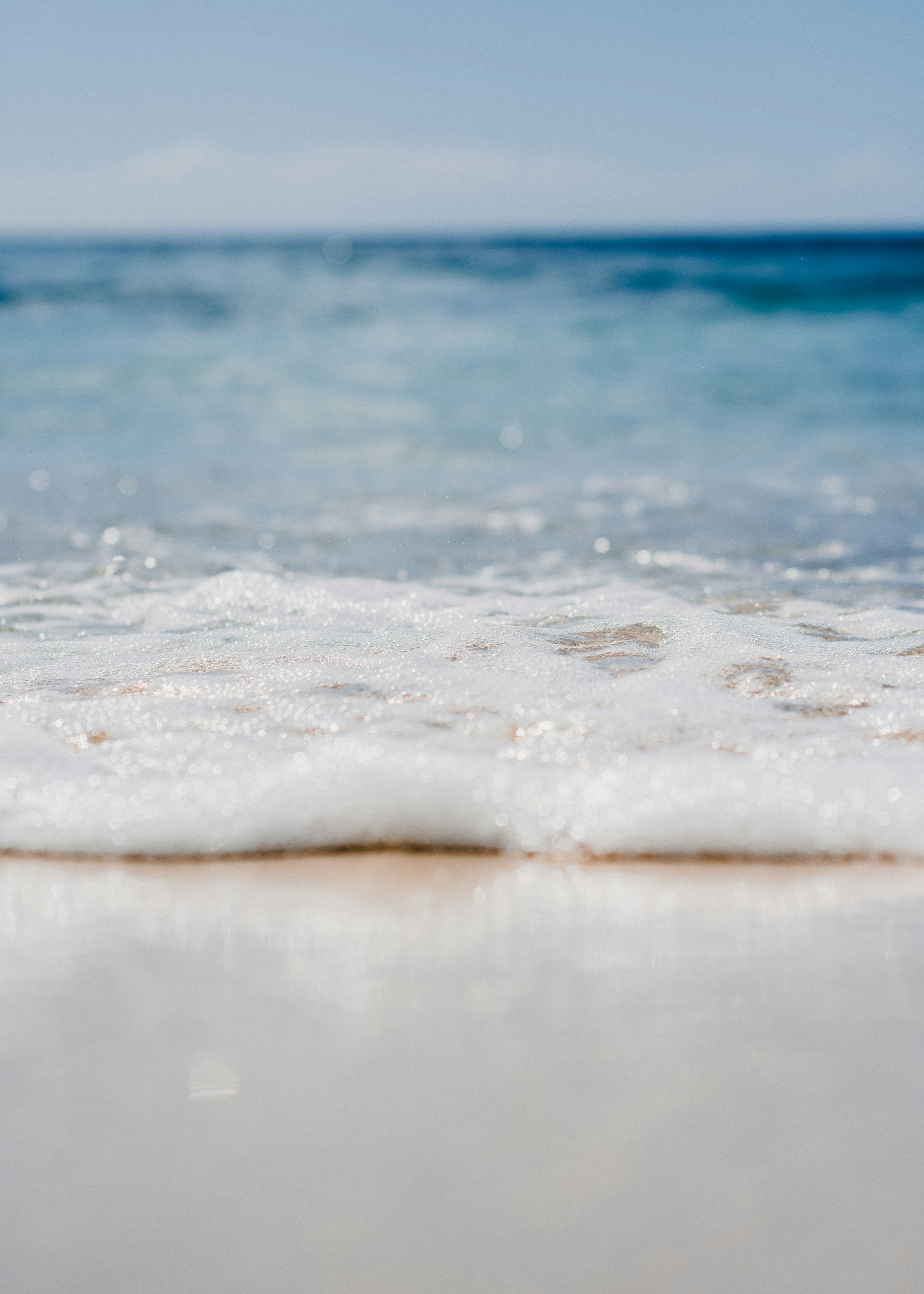 Close-up view of gentle ocean waves washing onto a sandy beach under a clear sky, reminiscent of the smooth curves and shimmer found in handcrafted gemstone jewelry.