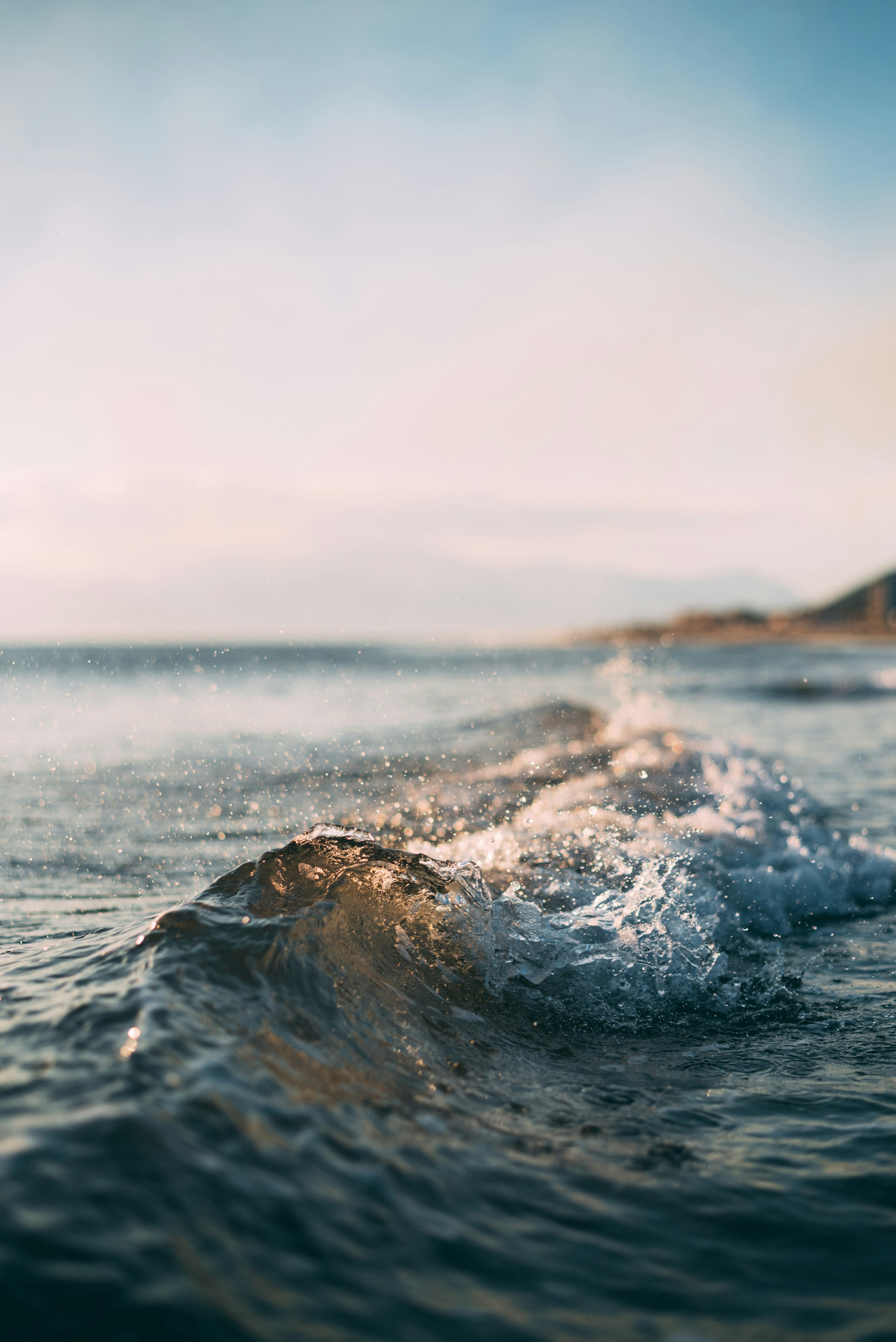 Close-up of a small ocean wave breaking near the shore with distant mountains in the background under a soft, pastel sky, resembling the smooth curves of handcrafted jewelry.